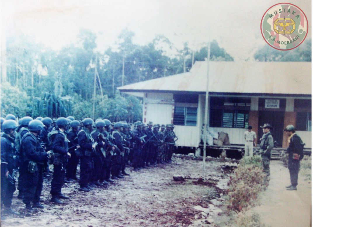 Dozens of uniformed soldiers line up in front of a wooden building during a briefing